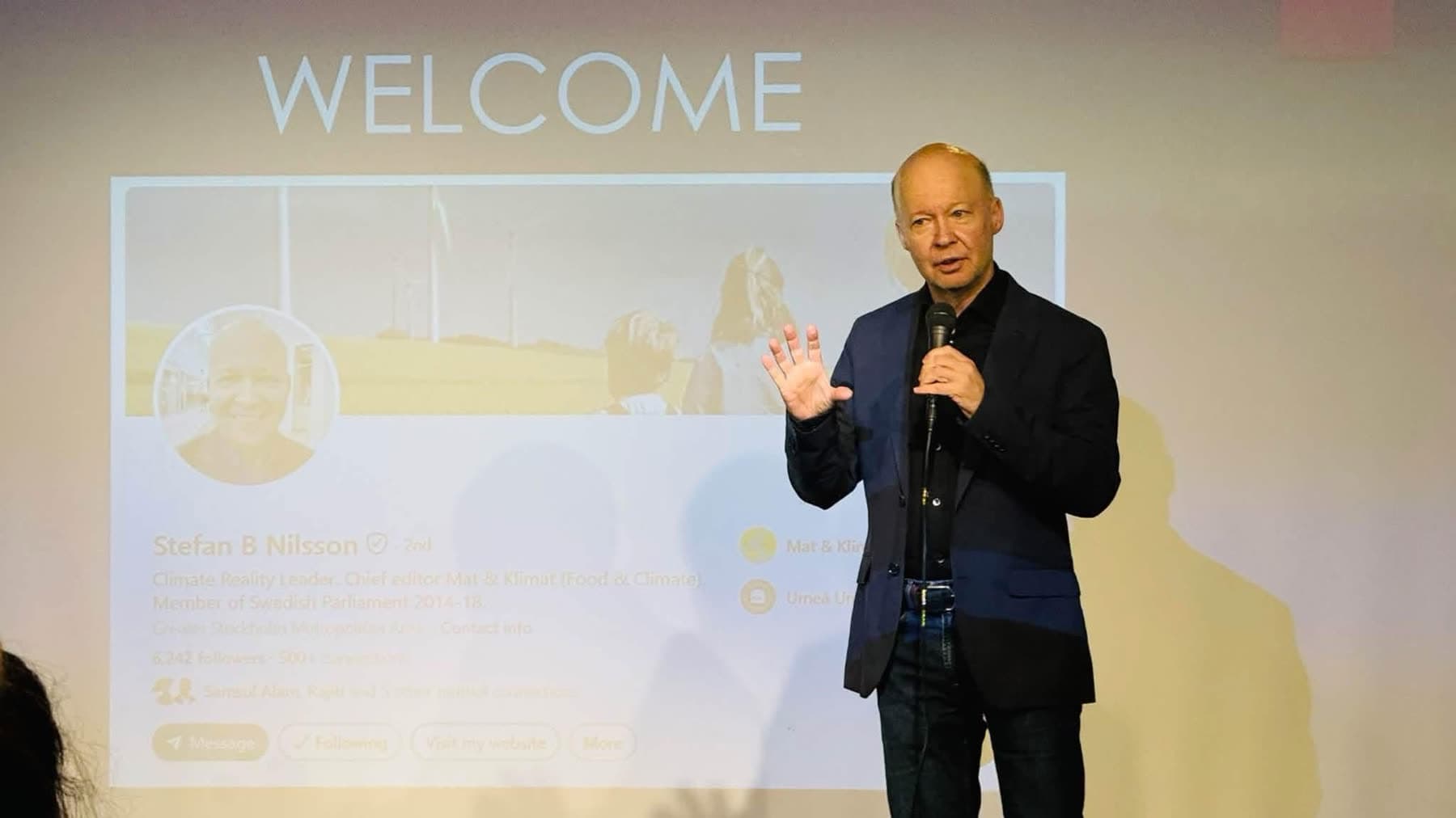 A speaker welcomes the audience with open arms in front of a welcome slide